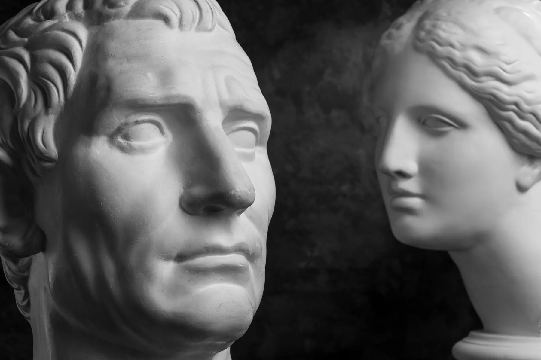 Two classical plaster busts photographed against a dark ground, a Roman male head and a female head, both seen near profile.